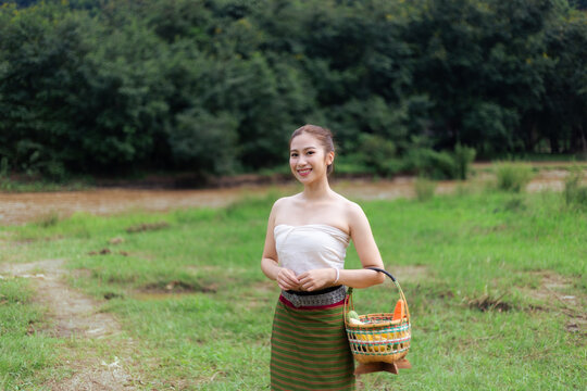 Young Asian woman wearing traditional clothing and carrying woven basket while standing in green rural field, expressing calm happiness during outdoor cultural scene