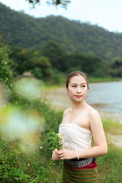 Asian woman holding wildflower near quiet lake and green mountain, wearing traditional clothing with calm expression in peaceful outdoor setting