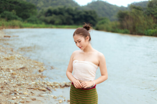 Young Asian woman in traditional clothing standing beside calm river with peaceful expression and lush forest landscape in background