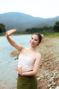 Smiling young Asian woman in traditional clothing posing beside mountain lake, graceful hand gesture, calm outdoor travel portrait with natural landscape background