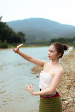 Young Asian woman in traditional clothing making graceful gesture beside calm lake and mountain landscape, creating serene cultural travel scene with natural beauty