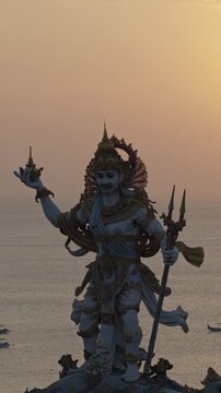Golden hour sunset over the Bali ocean horizon behind Dewa Baruna Hindu god statue with traditional outrigger boats jukung under warm orange sky Indonesia vertical aerial drone footage
