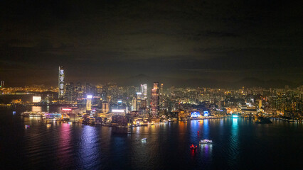 Fototapeta premium Aerial drone view of the Tsim Sha Tsui skyline in Hong Kong at night, neon waterfront buildings glowing across the black water of Victoria Harbour with boats passing