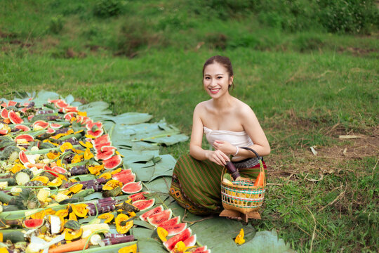 Smiling Asian woman wearing traditional clothing beside colorful fruit display and woven basket in rural green field, expressing joy during outdoor cultural market scene