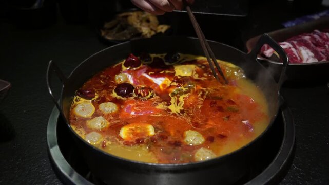 Close up of chopsticks holding raw beef roll into boiling spicy hot pot broth
