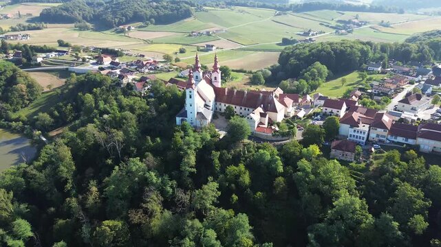 Aerial drone view of Holy Trinity Franciscan Monastery in Sveta Trojica, Slovenia, featuring three bell towers, historic red rooftops and the nearby Trojica Lake.