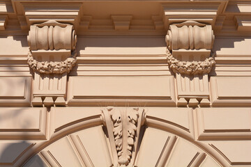 A detailed close-up of a beige building facade featuring a decorative acanthus leaf keystone above an arch, beneath two ornate corbels with hanging floral garlands. © nahhan