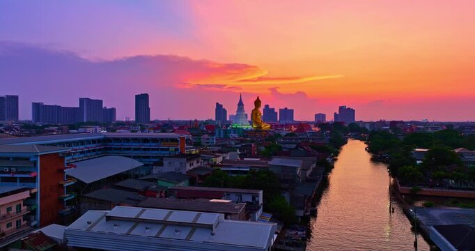 A breathtaking sunset over Bangkok, where the golden Buddha at Wat Paknam Phasi Charoen glows warmly against a dramatic twilight sky, with reflections shimmering across the peaceful canal below.