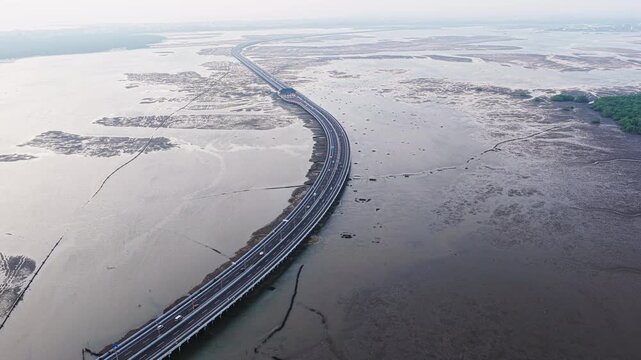 Traffic on a coastal causeway cars travel a long elevated highway over exposed tidal landscape and shallow water in soft hazy light aerial drone footage