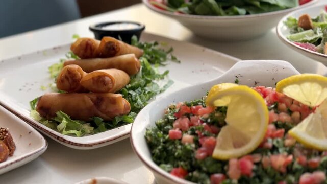 Close up of Middle Eastern appetizers including fried spring rolls and fresh tabbouleh salad with lemon slices. The food is served on white plates at a restaurant table during daytime.