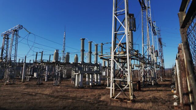 An outdoor electrical substation with metal towers, transformers, and insulators stands on dry brown grass under a clear blue sky, while shadows lengthen across the ground as the sun moves.