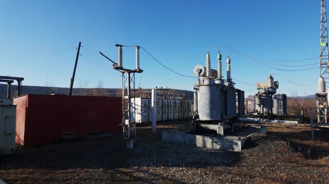 Large electrical transformers with colorful insulators stand on gravel ground within an outdoor substation under a clear blue sky with distant hills.