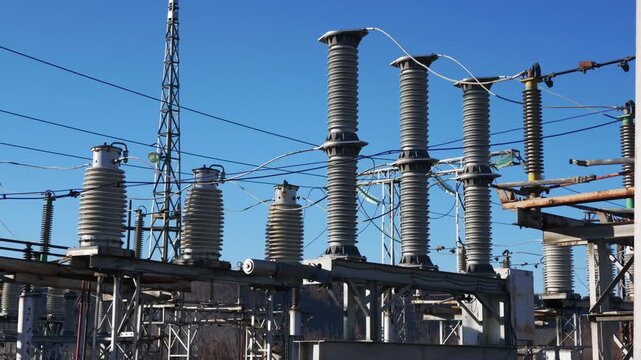 A slow right pan reveals a power substation with weathered cylindrical insulators on metal frameworks, connecting wires, and a tall transmission tower against a clear blue sky with distant hills.