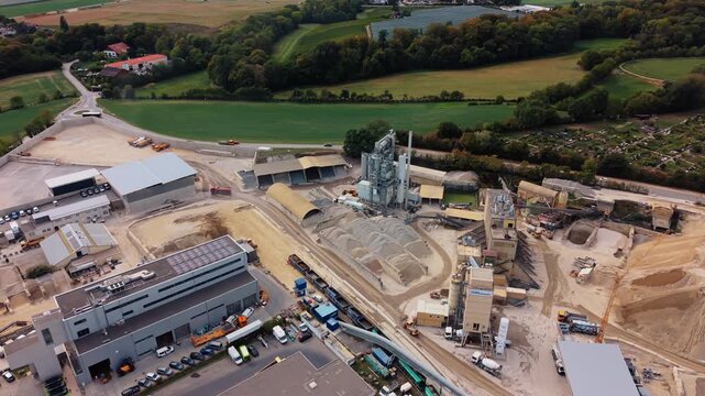 A drone turns above La Ballastiere in Gland, Vaud, Switzerland, showing aggregate stockpiles, processing towers and industrial buildings bordered by fields and trees.