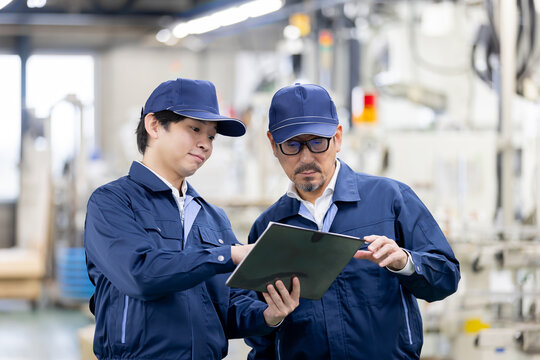 製造工場でタブレット端末を見ながら作業内容を確認し、打ち合わせをする日本人の若手とベテランの男性作業員2人