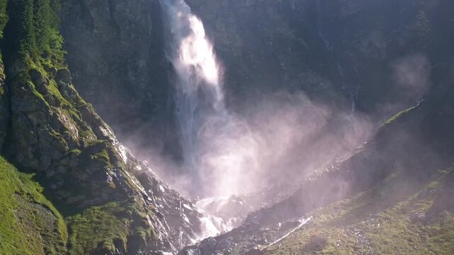 Waterfall plunging down a dark rocky cliff. Waterfall surrounded by dense sunlit mist. Powerful waterfall cascading over rugged rocks. Towering waterfall with white water spray.