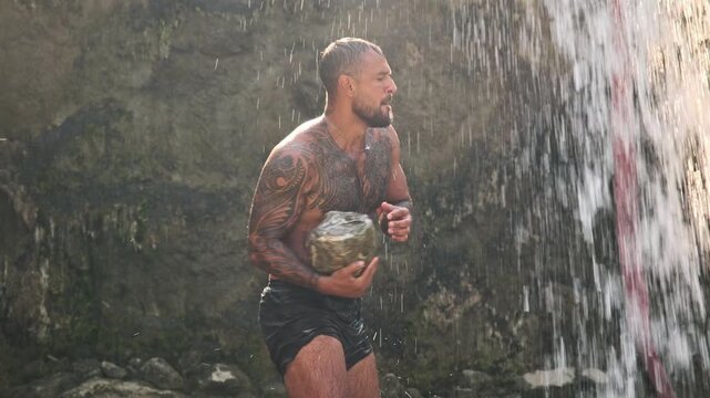 Muscular tattooed man holding heavy stone under waterfall during workout. Strong athletic man with tattoos training with a rock near waterfall. Man doing strength training in nature under splashes.