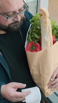 Surprised plus size man with shopping paper bag full of food looks at paper receipt on escalator in supermarket. Male shocked by food inflation and rising prices in shopping mall