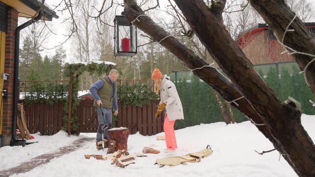 A man splitting firewood with an axe on a tree stump in a snowy yard in Finland, while a woman watches nearby during a winter day