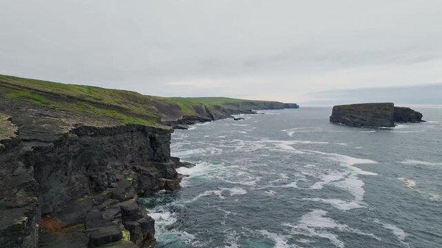 An offshore sea stack rises beyond rocky sea cliffs on the Kilkee coast in County Clare, Ireland, with Atlantic surf and green headlands in an aerial destination view.