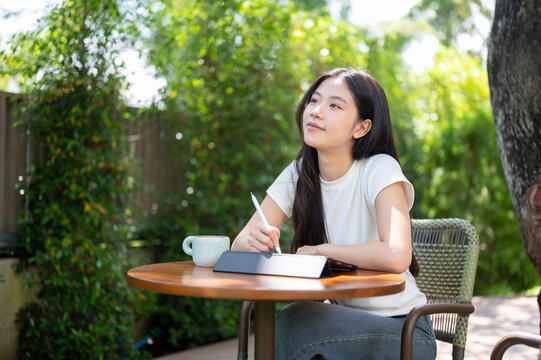 Asian Woman Holding Stylus and Daydreaming at Garden Cafe Table