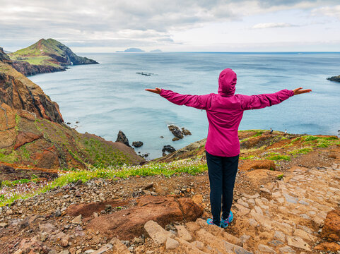 A tourist in a pink jacket standing with open arms on a mountain peak enjoying the ocean view at Ponta de Sao Lourenco, Madeira, Portugal.