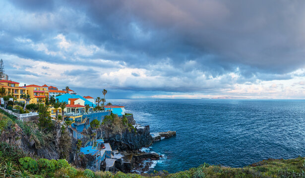 Colorful residential houses on a coastal hill looking over a dramatic dark stormy ocean under a cloudy sky in Canico, Madeira, Portugal.