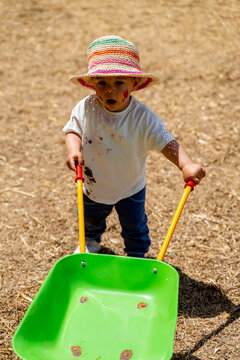 White child playing with toy wheelbarrow outdoors on straw ground