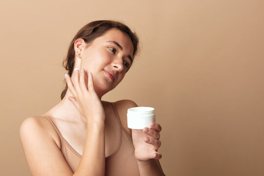 Cosmetics private label, young woman holds white unlabeled jar and touches her cheek on a beige background. Concept of skincare branding, beauty packaging mockup, and natural self care.