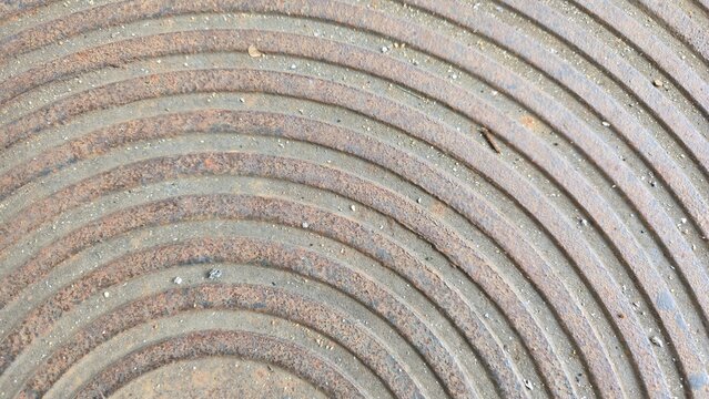 Old cast iron manhole cover with concentric circular pattern. Grunge industrial metal background with curved lines. Close-up of weathered metallic manhole texture showing circular ridges and rust.
