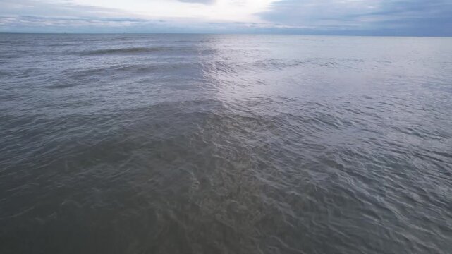 Aerial view of textured ocean water approaching a dark sandy coastline, with fine wave ripples and reflected light across the sea, in a natural frame.