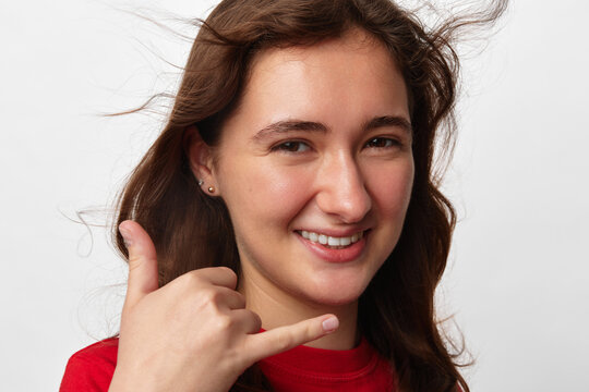Customer support agent, smiling young woman with long brown hair in red shirt making a call me gesture with her hand. Close-up portrait on a white background. Concept of contact, communication and