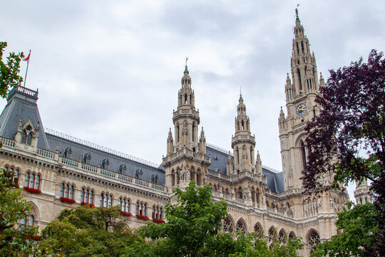Detailed low angle shot of historic Vienna City Hall featuring ornate Gothic revival architecture, tall spires, clock tower, and surrounding trees under overcast sky.