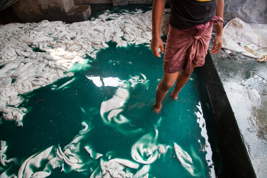 Barefoot worker stands in a turquoise tannery vat full of white wet hides under sunlight in a concrete hall, Hazaribagh, Bangladesh