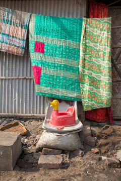 Colorful quilts hanging on a corrugated metal wall above a red plastic duck potty and bricks outside a tannery shack, Hazaribagh, Bangladesh