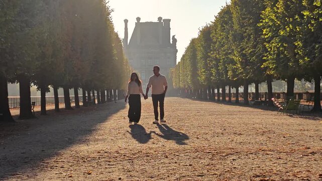 In the beautiful Tuileries Garden in Paris, France, a couple strolls hand in hand, basking in the warm sunlight and enjoying the romantic atmosphere. Trees line their path, creating a serene backdrop.