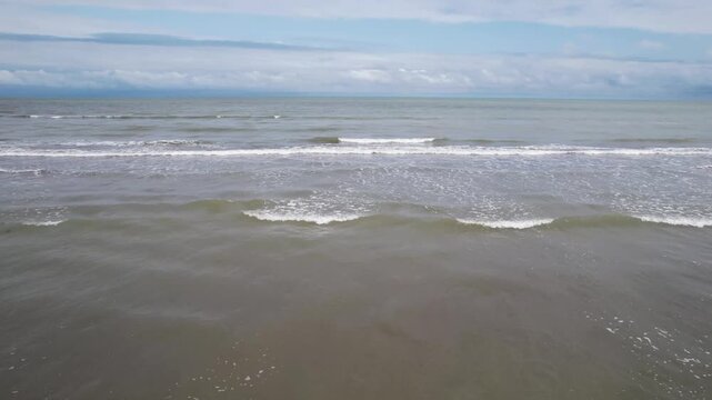 Small parallel wave crests moving toward the coast across a calm sea, beneath a diffuse gray sky and distant shoreline, in a detailed wide angle view.