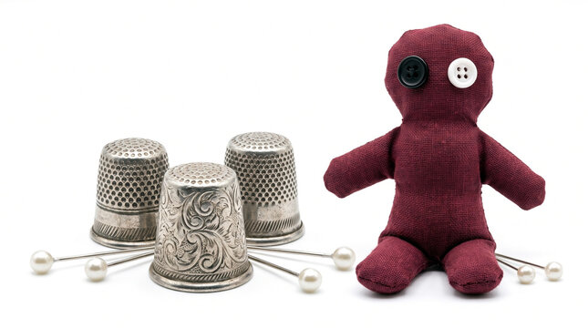 A handmade maroon rag doll with button eyes sits next to vintage silver thimbles and pearl-headed pins on a white background