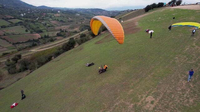 Orange paragliding canopy near a grassy hillside with people on the ground, seen against the fields and roads of a rural valley, as viewed from above.