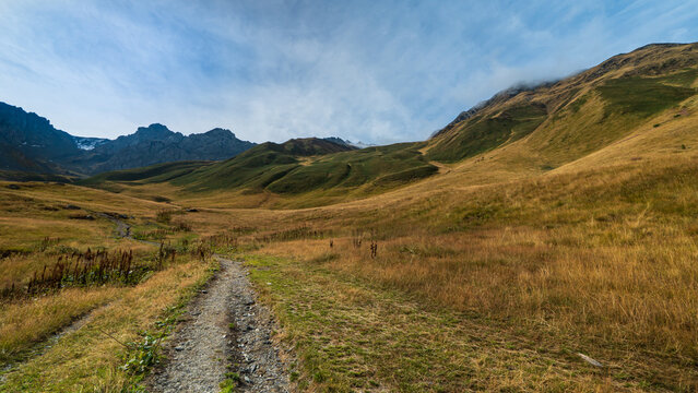 Hiking trail crossing a wide alpine grassland surrounded by rugged mountains near Juta, Georgia