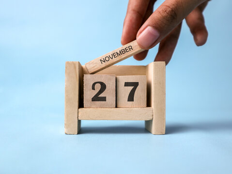 A woman's hand holds a wooden block with November written on it on a wooden block calendar for November 27th against a light blue background.