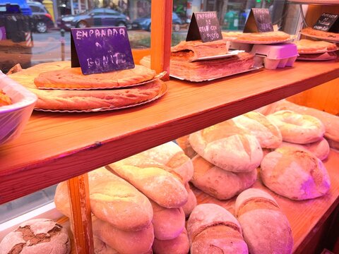 View of a traditional bakery display case showing a variety of rustic breads, loaves, baguettes, and freshly baked homemade empanadas with small descriptive chalkboard signs.