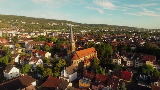 A drone pulls back from a church with a pointed clock tower and orange tiled roof in Munich, Germany, revealing surrounding houses, trees and the wider cityscape.