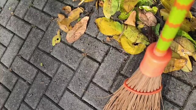 Close-up of a traditional broom sweeping fallen leaves and debris from a brick pavement during outdoor cleaning and maintenance.