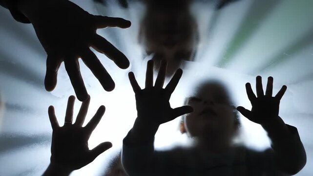 Silhouettes of children's hands pressing against a translucent glass pane with bright backlighting