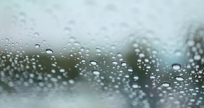 Raindrops on a car windshield with blurred green trees in the background, rainy weather, wet driving conditions, reduced visibility and travel in the rain, close-up