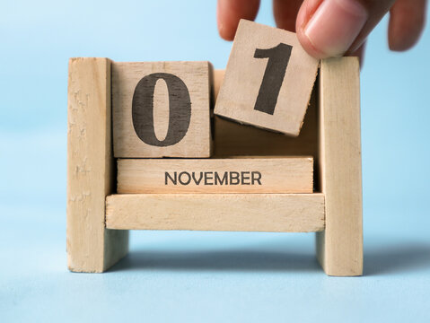 A woman's hand is holding a wooden block with the number 1 written on it on a wooden block calendar for November 1st on a light blue background.