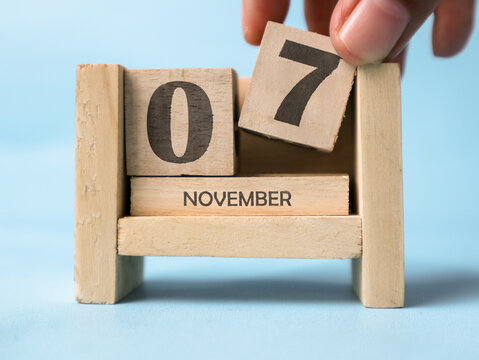 A woman's hand is holding a wooden block with the number 7 written on it on a wooden block calendar for November 7th on a light blue background.