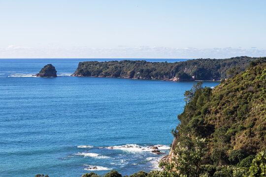 A rugged forested headland rises above the deep blue ocean in Coromandel, with a small rocky island and clear summer sky completing the serene coastal scene