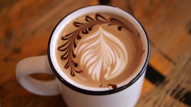 Overhead view of cappuccino with intricate leaf latte art in white ceramic cup. Foamed milk forms feathered heart pattern across rich coffee, resting on warm wooden cafe table, filling cup to rim.